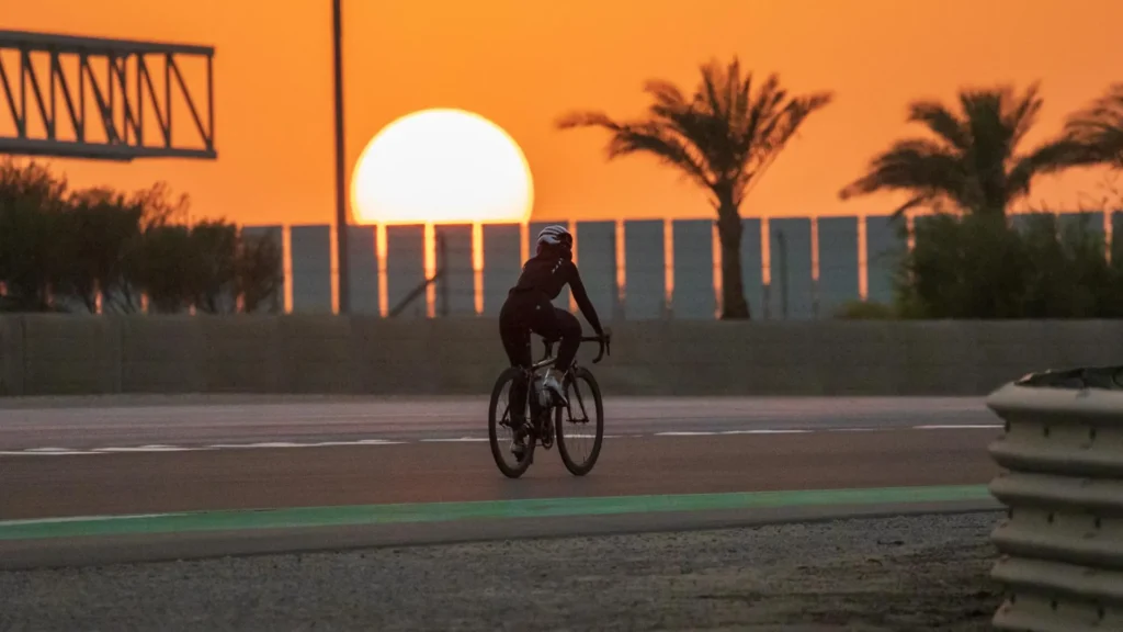 A silhouetted triathlete cycles on a closed track during an early morning ride, with the bright orange sun rising directly behind a line of palm trees and a fence. The atmosphere suggests training for the Dubai T-100 Triathlon.